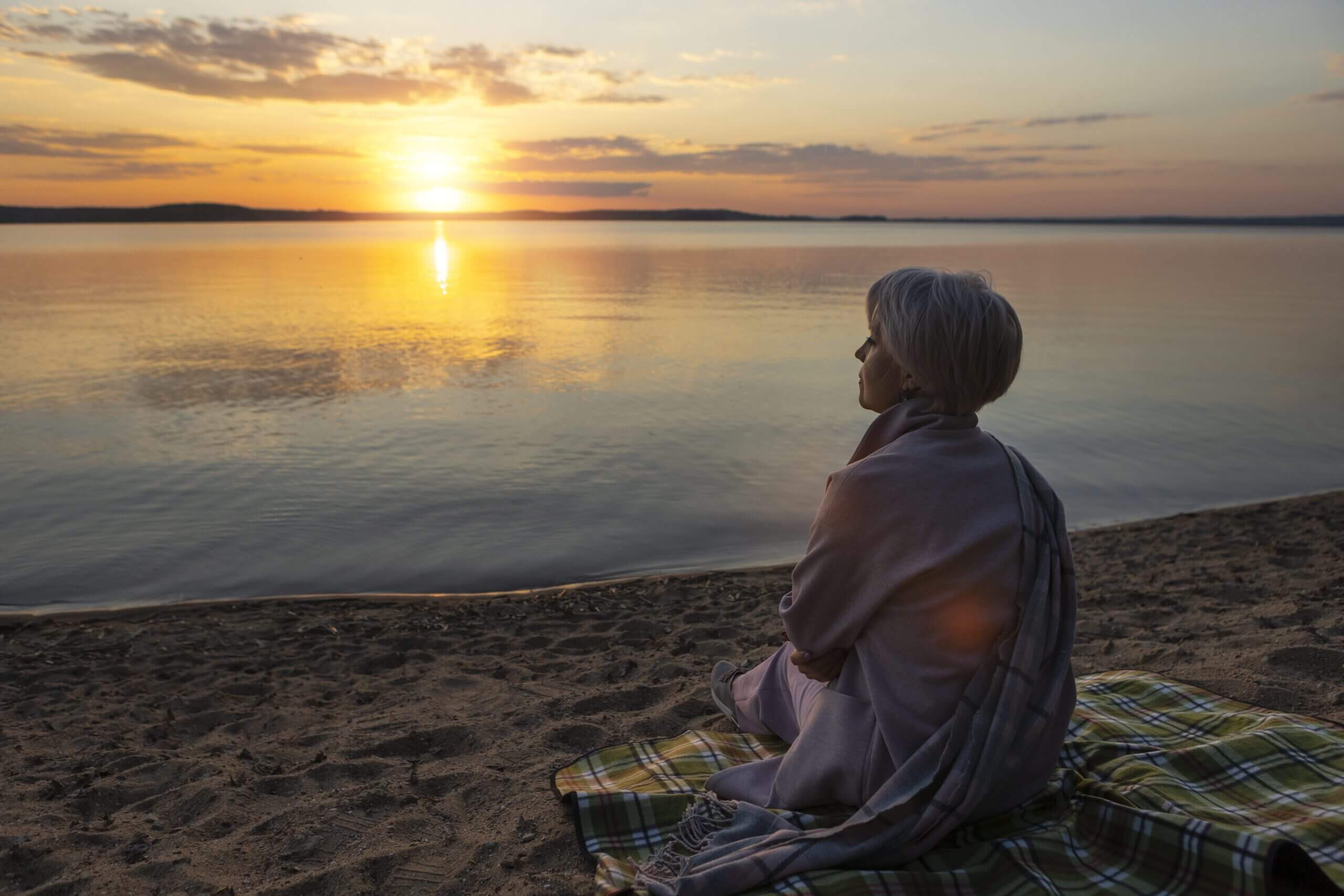 Una imagen serena y emotiva de una mujer sentada al borde de un acantilado al amanecer, envuelta en un chal ligero, mirando al horizonte con una expresión apacible e introspectiva. Una suave luz dorada baña el paisaje, simbolizando nuevos comienzos y claridad interior. El entorno se siente sagrado y abierto, con sutiles elementos naturales como flores silvestres o una suave brisa. El tono es cálido, conmovedor y profundamente humano: una metáfora visual de la sanación, la transformación y el poder sereno de estar acompañado en viajes emocionales.